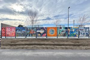 Bunte Wandbilder auf einer Lärmschutzwand am Cottbuser Hauptbahnhof zeigen Motive zur Mobilität von morgen neben einer Straße unter wolkigem Himmel.