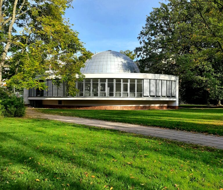 Rundes Planetarium Cottbus mit silberner Kuppel zwischen hohen Bäumen und grüner Rasenfläche im sonnigen Park.