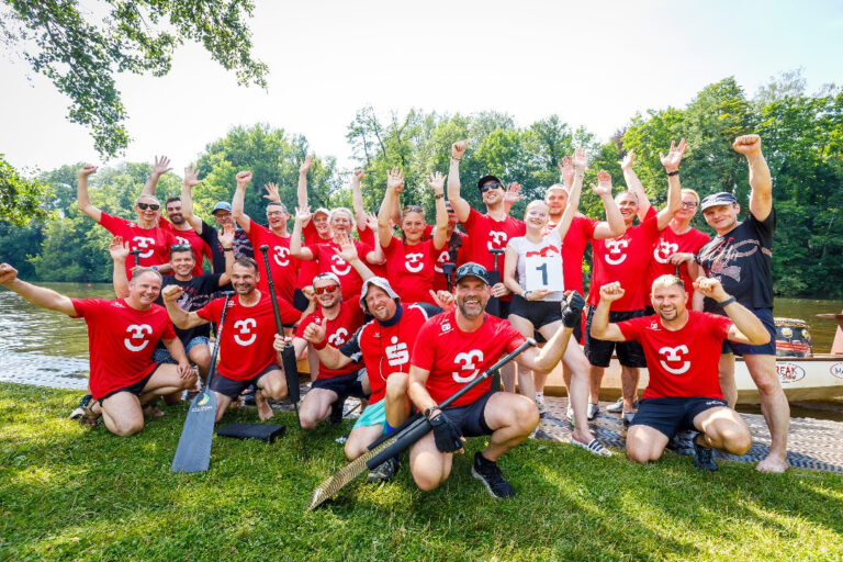 Gruppe in roten Shirts jubelt am Ufer der Spree, einige mit Paddeln, nach einer Drachenbootregatta.
