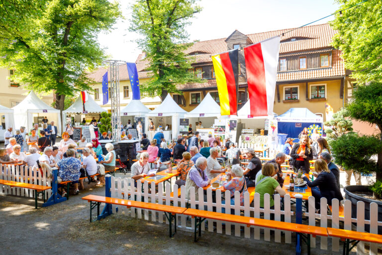 Beim deutsch-polnischen Bürgerfest auf dem Klosterplatz sitzen Gäste zwischen Marktständen, Fahnen von Deutschland, Polen und der EU unter Bäumen.