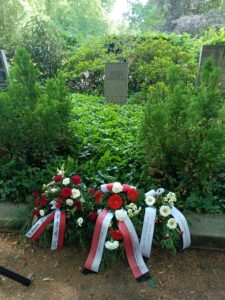 Am Grab von Gustav Schuft im grünen Friedhofsgehölz liegen mehrere Kränze und Blumen mit Schleifen vor dem schlichten Grabstein zum 150. Geburtstag.