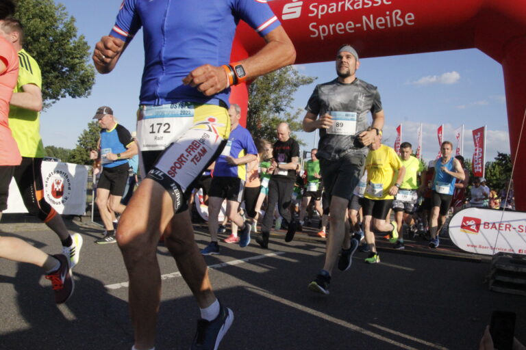 Beim Laufstart bei den Ostsee-Sportspielen in Cottbus/Chóśebuz starten zahlreiche Läufer unter dem roten Zielbogen und tragen Startnummern.