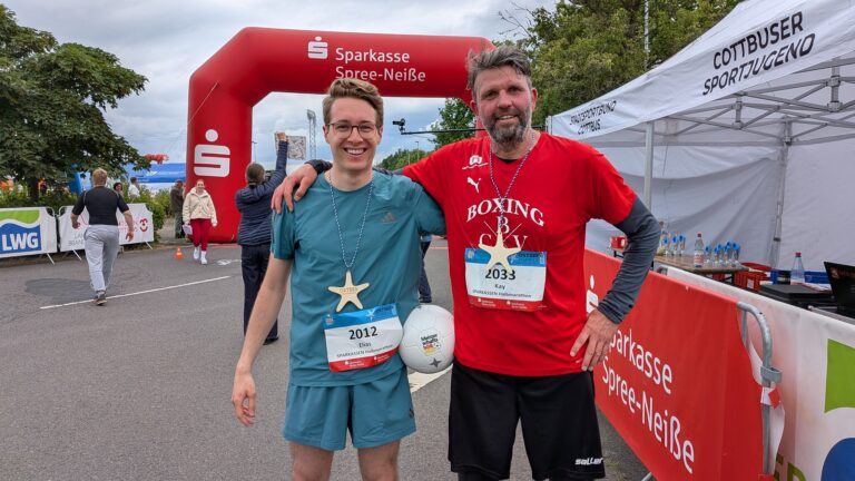 Zwei Männer mit Startnummern stehen lächelnd vor einem roten Sparkasse-Spree-Neiße-Startbogen beim Lauf.