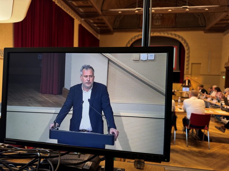 Im Ratssaal spricht Tobias Schick an einem Rednerpult, während Dan Bishop auf dem Monitor und die Stadtverordnetenversammlung im Hintergrund zu sehen sind.