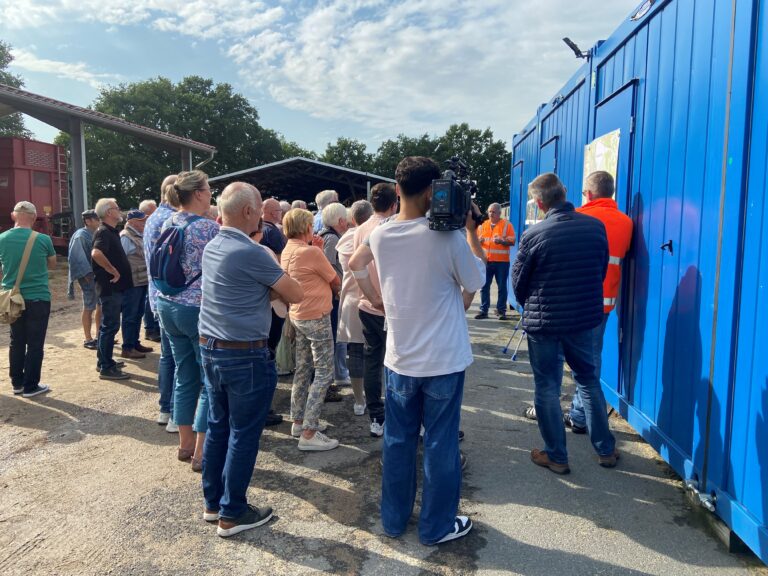 Menschen stehen vor einem blauen Baustellencontainer und hören einem Mann in Warnweste zu, während ein Kameramann filmt.
