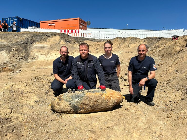Vier KMBD-Mitarbeitende knien neben einer großen Fliegerbombe in einer Sandgrube; dahinter stehen Container und Absperrungen am Bahnhof Cottbus.