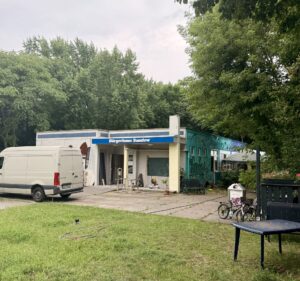 Bürgerhaus Sandow in Sandow mit blauem Schriftzug, davor ein weißer Transporter, Fahrräder und ein Gartenbereich am Rand eines dichten Waldstücks.