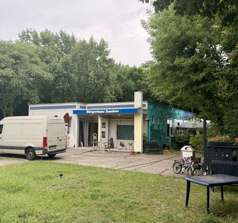 Bürgerhaus Sandow in Sandow mit blauem Schriftzug, davor ein weißer Transporter, Fahrräder und ein Gartenbereich am Rand eines dichten Waldstücks.