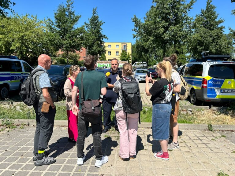 Journalisten mit Mikrofonen befragen einen Mann zwischen Polizeiwagen auf einem Parkplatz in Cottbus, während er vor laufenden Kameras spricht.