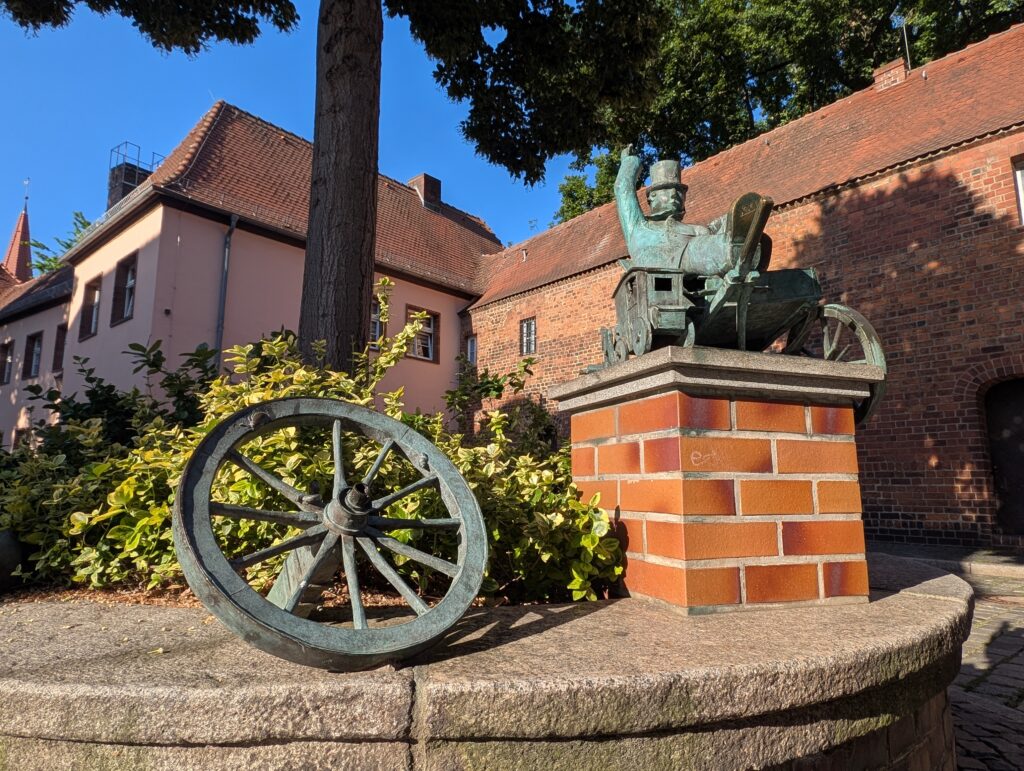 Bronzene Statue eines Mannes auf einem Wagen neben einem großen Speichenrad vor Backsteinmauern und roten Dächern.