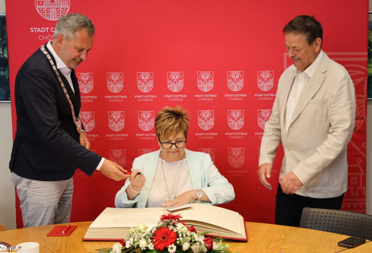 Dr. Cornelia Schmidt trägt sich am Tisch in das Goldene Buch der Stadt Cottbus/Chóśebuz ein, begleitet von Oberbürgermeister Tobias Schick und einem weiteren Mann.