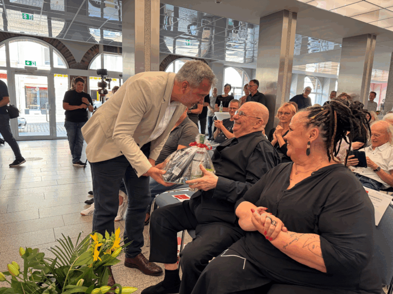 Im Rathausfoyer überreicht ein Mann im hellen Sakko einem sitzenden Gast ein verpacktes Geschenk, während Gäste zusehen.