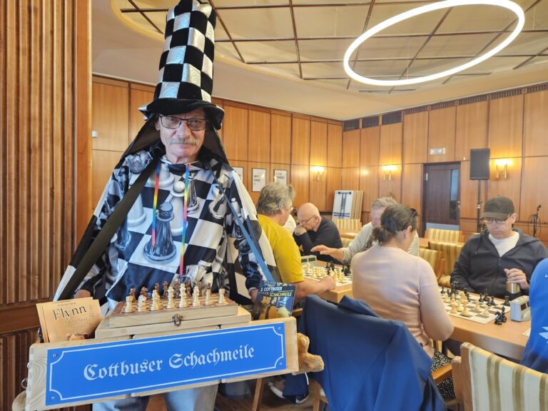 Eine kostümierte Person präsentiert ein Schachbrett mit dem Schild „Cottbuser Schachmeile“ in einem Raum voller Spieltische.