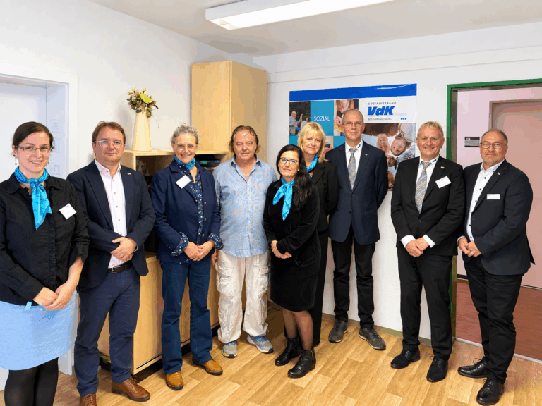 Acht Personen stehen für ein Gruppenfoto vor einem VdK-Plakat in den neuen Räumen