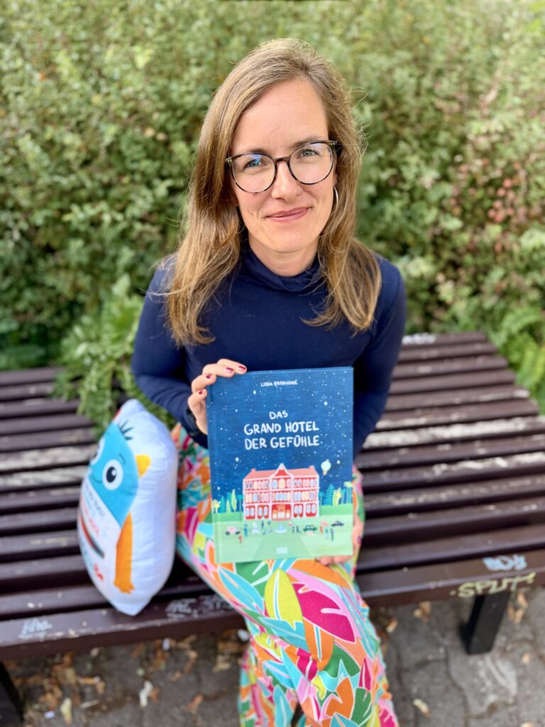 Frau sitzt auf Bank und hält das Buch Das Grand Hotel der Gefühle sowie ein Kissen