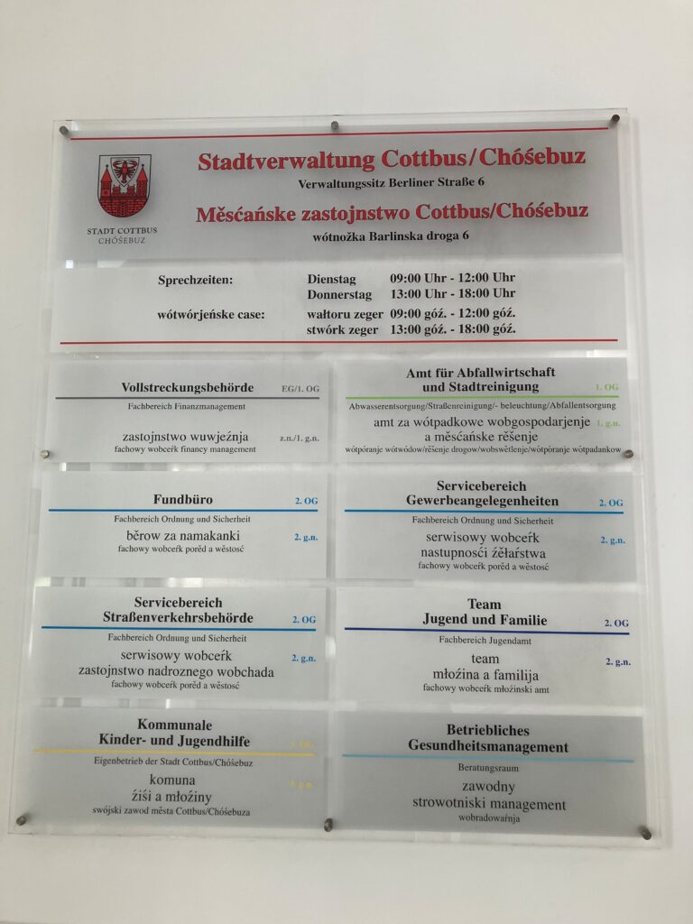Mehrsprachiges Hinweisschild der Stadtverwaltung Cottbus/Chóśebuz mit Abteilungen, Sprechzeiten und Raumangaben.