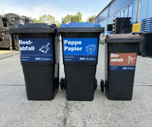 Vor dem Hof stehen Tonnen mit gelben, blauen und braunen Deckeln für Restabfall, Leichtverpackungen sowie Pappe und Papier.