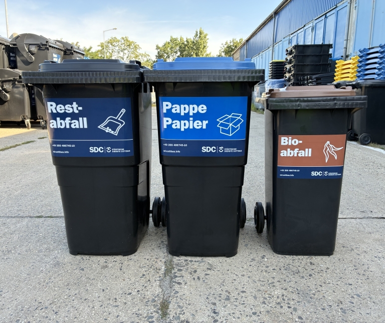 Vor dem Hof stehen Tonnen mit gelben, blauen und braunen Deckeln für Restabfall, Leichtverpackungen sowie Pappe und Papier.