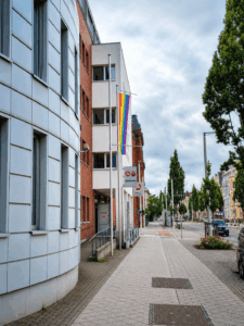 Vor dem Gebäude von Jobcenter und Agentur für Arbeit in Cottbus hängt eine Regenbogenflagge an der Straße neben dem Gehweg unter wolkigem Himmel.