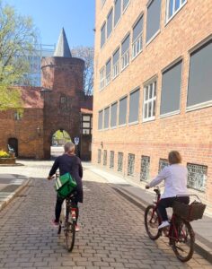 Zwei ältere Frauen fahren mit Fahrrädern durch einen historischen Torbogen in Cottbus/Chóśebuz, vorbei an Backsteinbauten der Altstadt bei Sonnenschein.