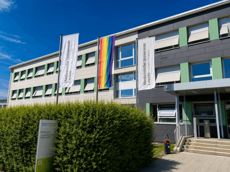 Vor dem MUL-CT hängen zwei Banner der Medizinischen Universität Lausitz – Carl Thiem neben einer Regenbogenflagge am modernen Klinikgebäude in Cottbus.