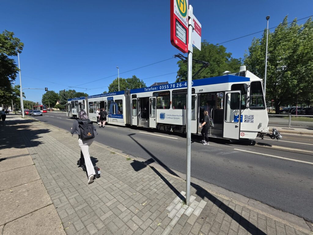An der Haltestelle Hüfelandstraße steigen Fahrgäste in eine Straßenbahn ein, während weitere Personen warten.