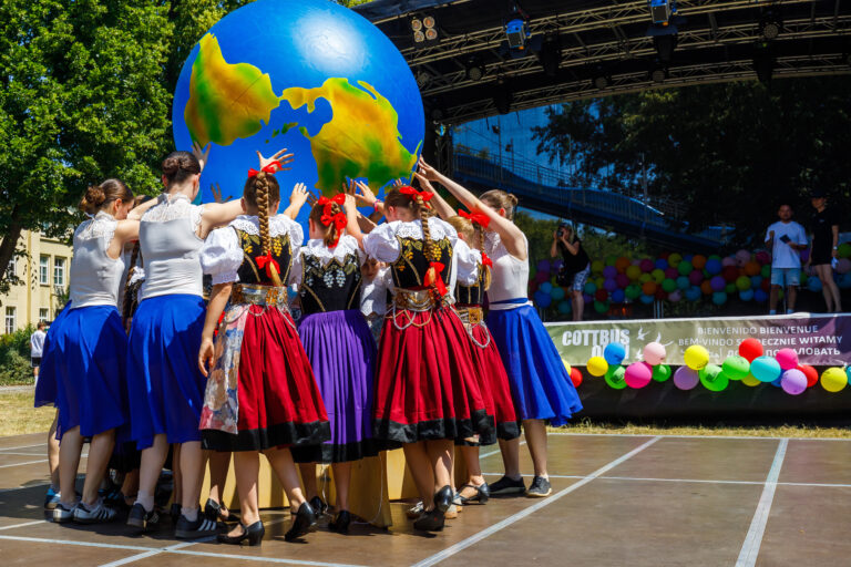 Mehrere Kinder in farbenfrohen Trachten stehen mit erhobenen Armen vor einem großen Globus auf einer Bühne.