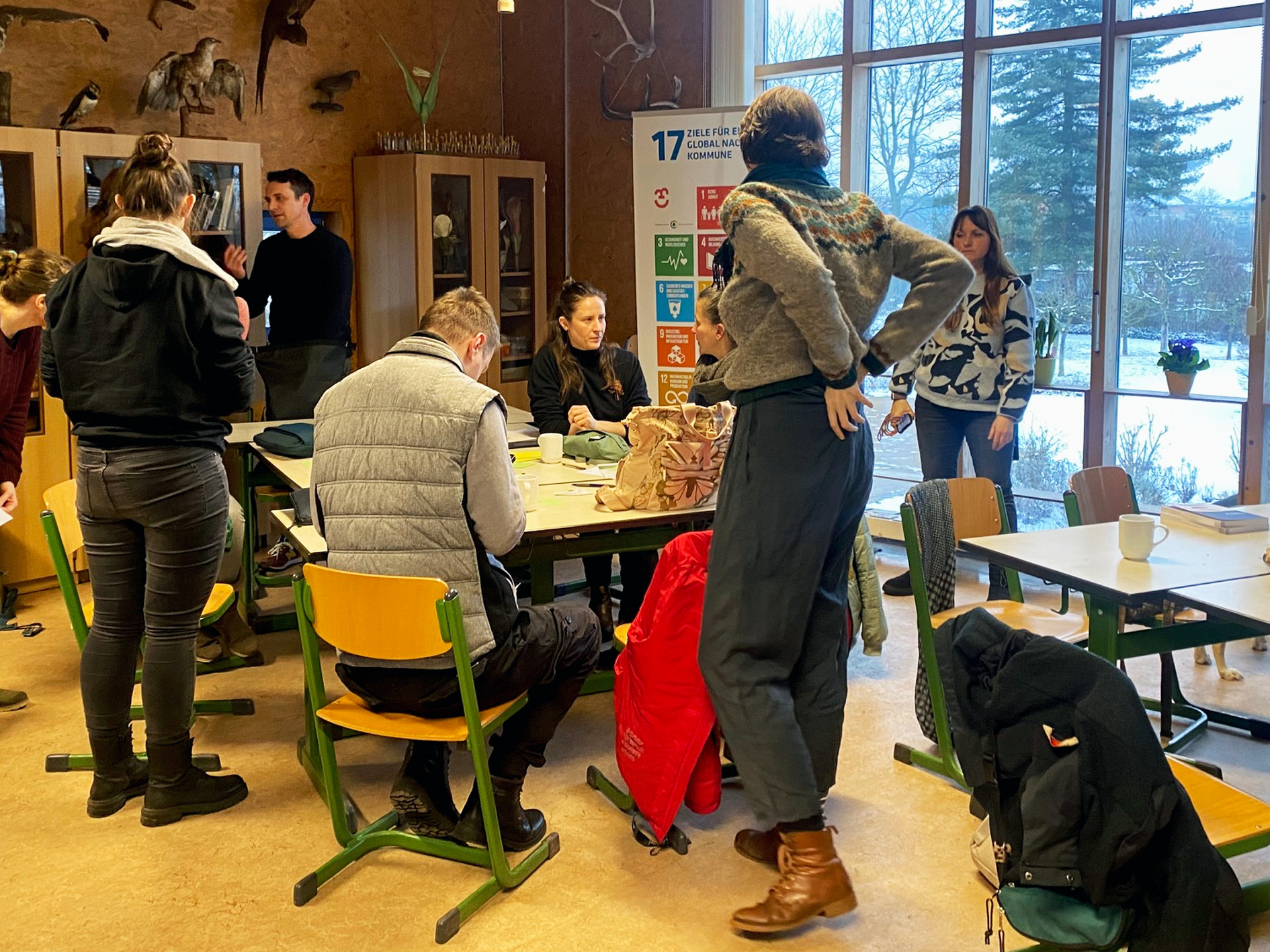 Menschen arbeiten in kleinen Gruppen und führen Gespräche. Im Hintergrund ist ein Roll-up mit den 17 Zielen der Bildung für nachhaltige Entwicklung (BNE) sichtbar.