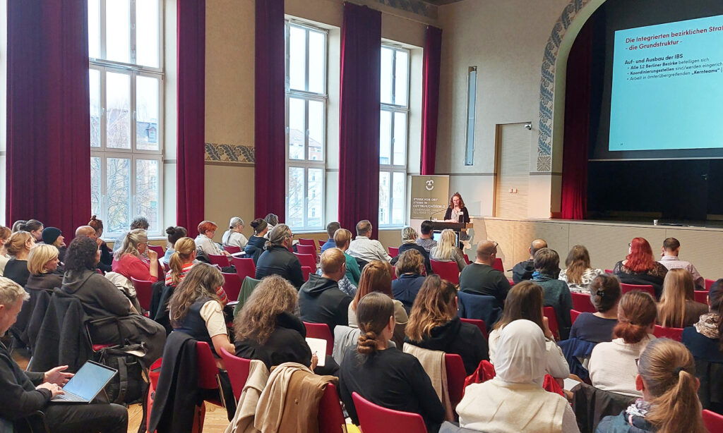 In einem Konferenzsaal sitzen mehrere Menschen in Reihen und hören aufmerksam zu, während eine Frau einen Vortrag hält.