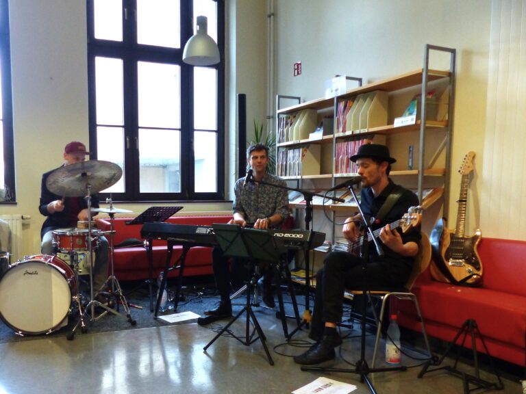 Eine Band spielt in der Bibliothek mit Schlagzeug, Keyboard und Bass vor Regalen und großen Fenstern bei einer 100-Jahre-Feier in Cottbus.
