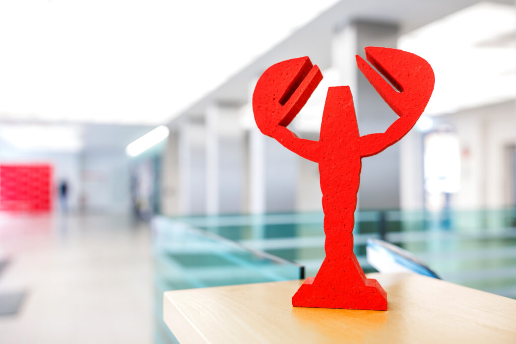 Ein rotes, stilisiertes Holzfiguren-Symbol steht auf einem hellen Tisch vor unscharfem Innenraum.