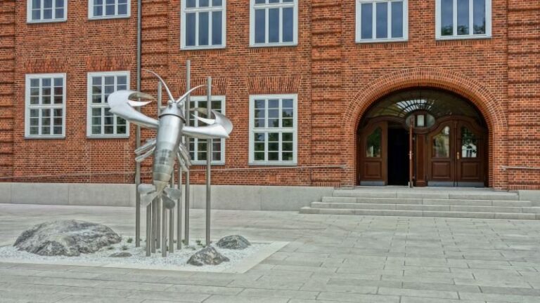 Vor dem roten Stadthaus in Cottbus/Chóśebuz steht eine silberne Skulptur, während der Eingangsbereich mit Bogen und Stufen sichtbar ist.