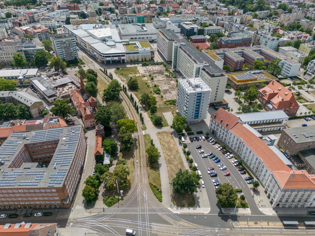 Blick über die Innenstadt von Cottbus/Chóśebuz mit Straßenbahnschienen, modernen Gebäuden, Parkflächen und grünen Baumbereichen im Stadtkern.