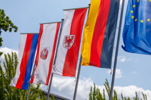 Mehrere Flaggen von Polen, Brandenburg, Cottbus und der EU wehen vor blauem Himmel mit etwas Grün.