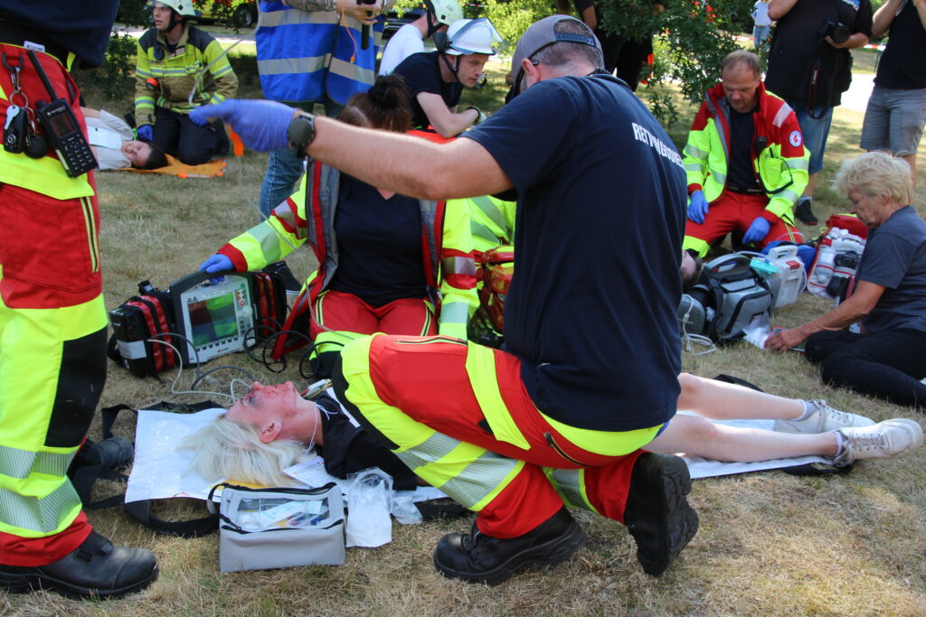Mehrere Einsatzkräfte versorgen auf einer Wiese verletzte Personen mit medizinischer Ausrüstung und Monitoren.