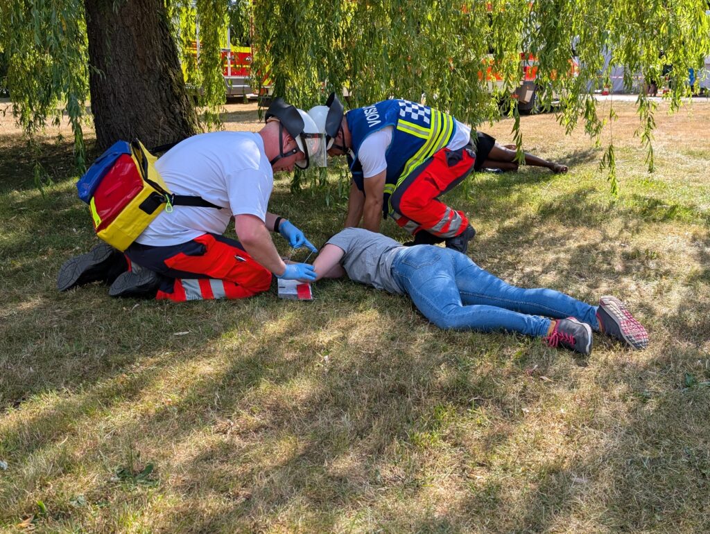 Zwei Rettungskräfte knien neben einer liegenden Person auf der Wiese und versorgen den Arm unter Weidenzweigen.