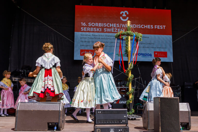 Mehrere Mädchen tanzen in sorbischer Tracht auf einer Bühne beim 16. Sorbischen/Wendischen Fest in Cottbus vor einem festlichen Hintergrund.