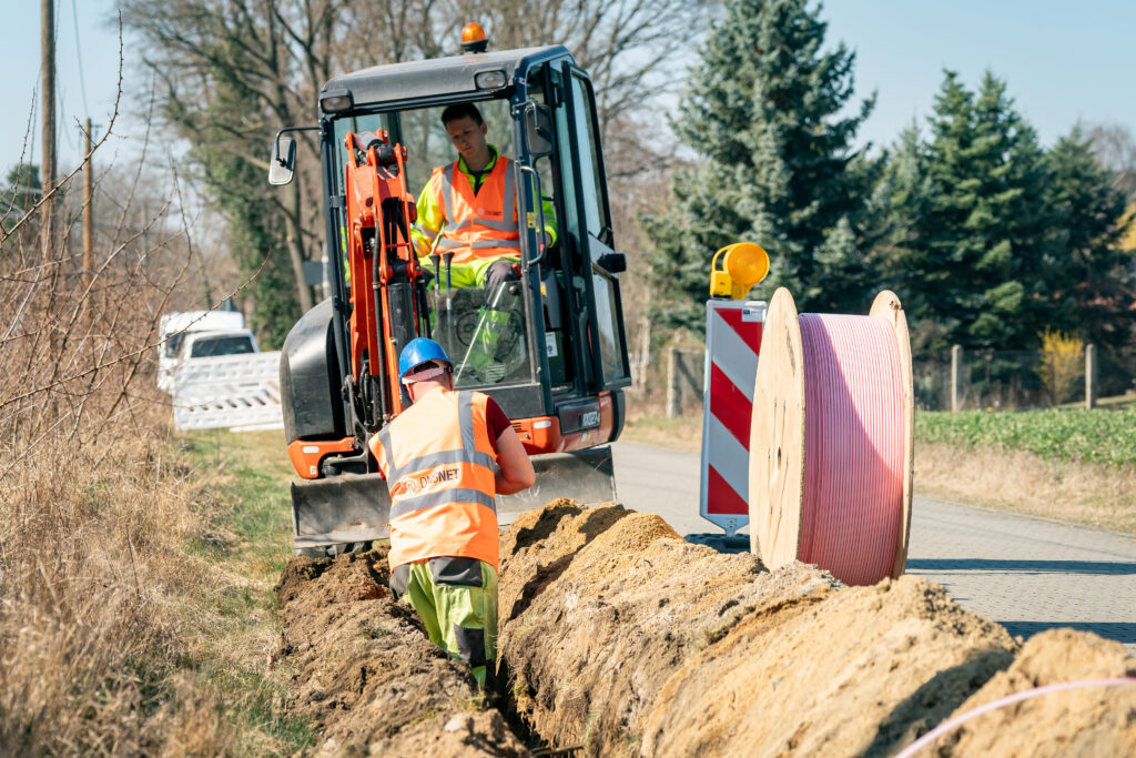 Beim Breitbandausbau in Cottbus/Chóśebuz verlegen Arbeiter mit Bagger und Kabeltrommel Glasfaserkabel in einem Straßengraben entlang der Landstraße.