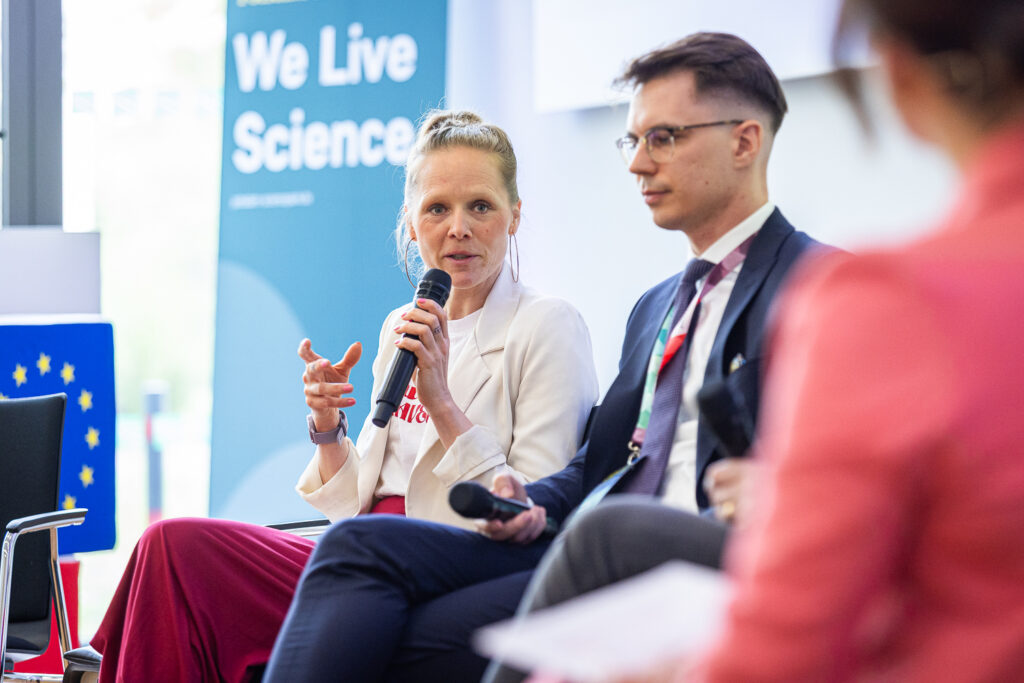 Dr. Maria Marquardt spricht mit Mikrofon auf einem Podium, neben ihr sitzt ein Mann im Anzug; im Hintergrund ein „We Live Science“-Banner und eine EU-Flagge.