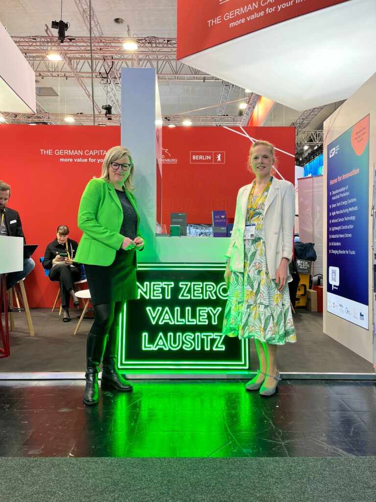 Martina Klement (links) und Dr. Maria Marquardt (rechts) stehen auf der Hannover Messe vor einem leuchtenden „Net Zero Valley Lausitz“-Schild. Im Hintergrund sind Messestände und Besucher zu sehen.