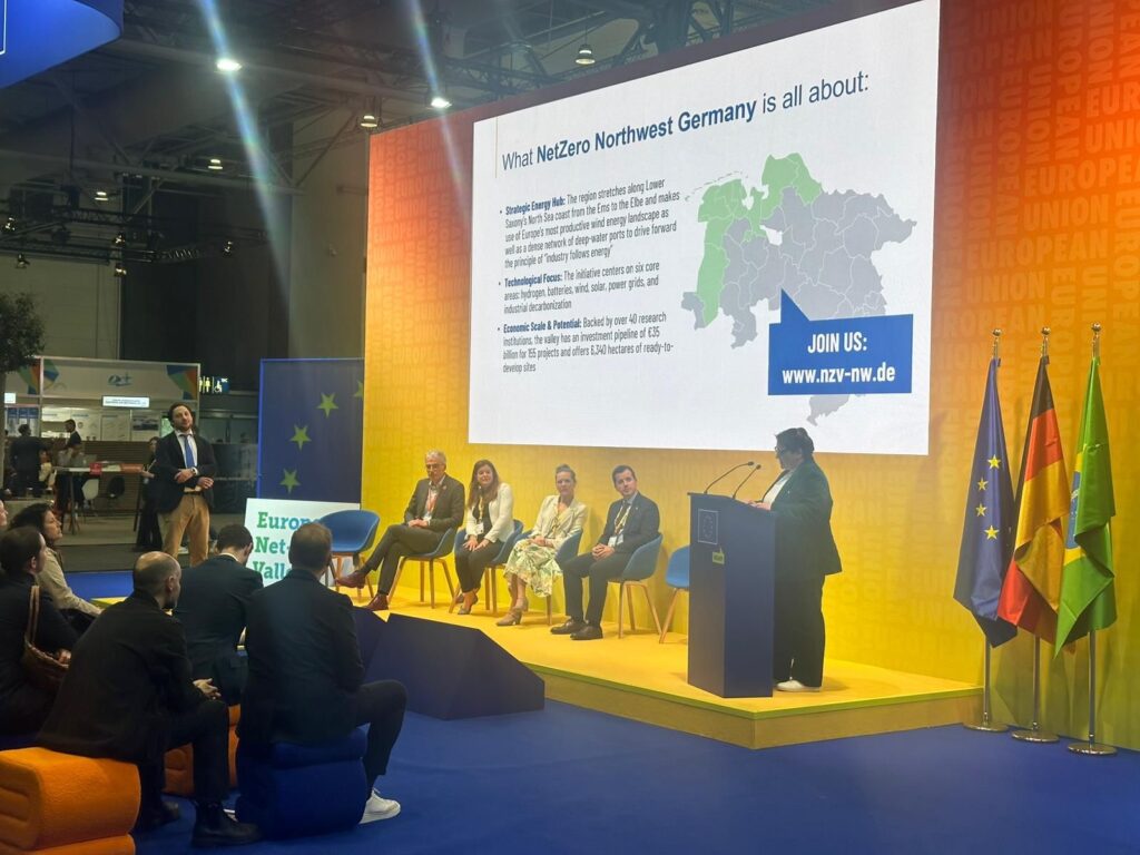 Podiumsdiskussion auf einer Messebühne: Mehrere Personen sitzen auf Stühlen, während eine Frau am Rednerpult spricht. Auf der großen Leinwand dahinter wird „Net Zero Northwest Germany“ mit Karte und Informationen präsentiert; davor sitzt ein Publikum.