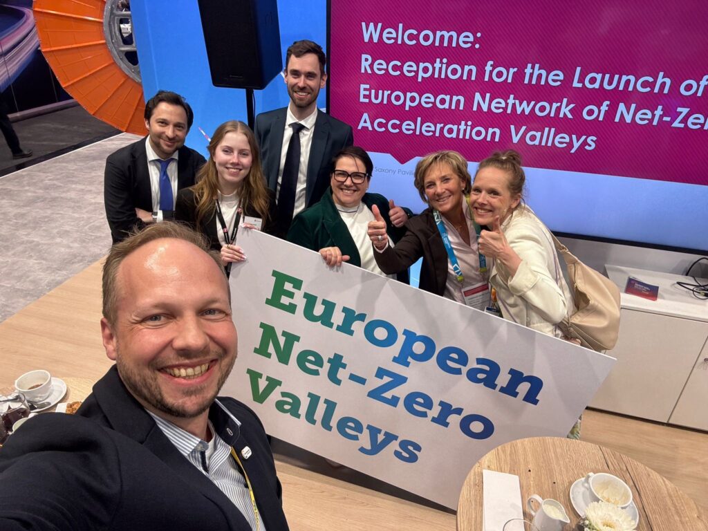 Gruppe von sieben Personen macht ein Selfie auf der Hannover Messe und hält ein Schild mit der Aufschrift „European Net-Zero Valleys“. Im Hintergrund ist ein Bildschirm mit einem Willkommenshinweis zur Auftaktveranstaltung des Netzwerks zu sehen.
