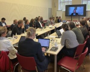 Teilnehmende eines Workshops sitzen an einem Konferenztisch, arbeiten an Laptops und verfolgen eine Präsentation, die per Videokonferenz auf einem Bildschirm zugeschaltet ist.