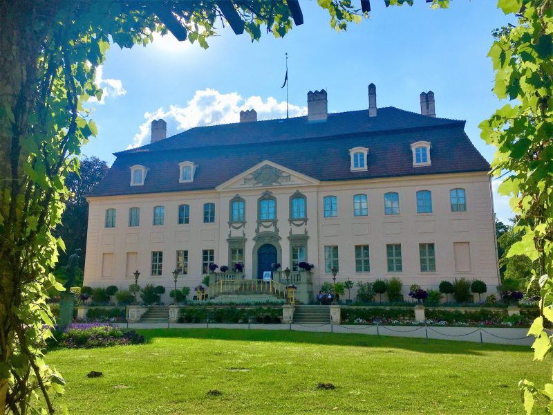 Umgeben von grünem Laub erhebt sich Schloss Branitz in Cottbus mit heller Fassade, dunklem Dach und gepflegter Gartenanlage unter blauem Himmel.