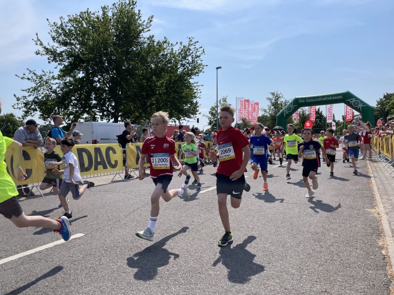 Kinder sprinten bei einem Straßenlauf unter einem grünen Zielbogen, während Zuschauer am Rand anfeuern und die Sonne hell über der Strecke steht.