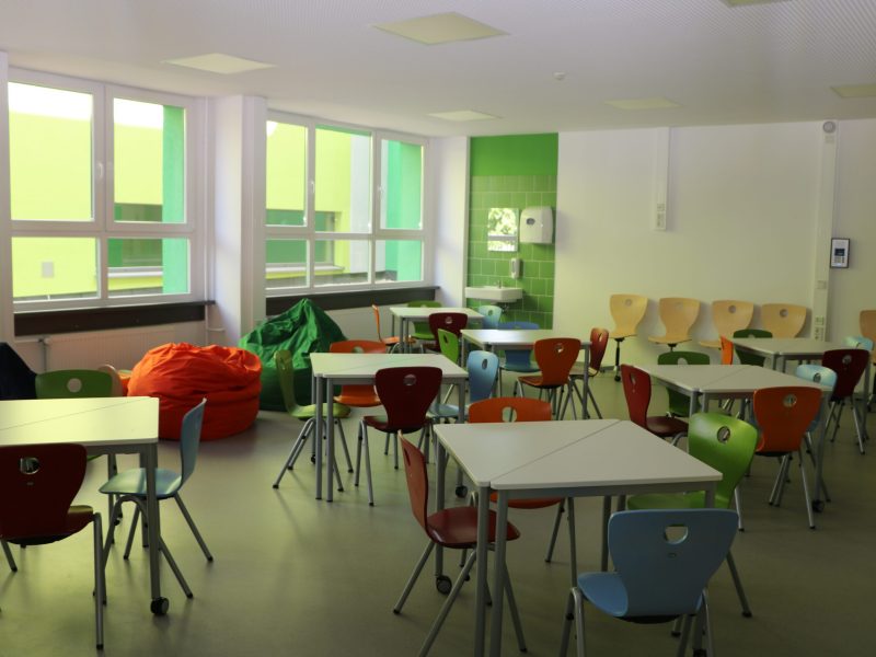 Ein heller Klassenraum mit bunten Stühlen, Tischen und Sitzsäcken, dazu große Fenster, grüne Wandfliesen und ein Waschbecken an der rechten Seite.
