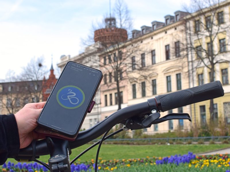 Eine Hand hält ein Smartphone am Fahrradlenker vor einem historischen Gebäude in Cottbus, während unten bunte Blumen den Parksaum säumen.