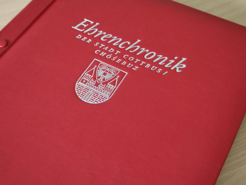 Ein rotes, lederartiges Buch mit dem Titel „Ehrenchronik der Stadt Cottbus/Chóśebuz“ und dem Stadtwappen liegt auf einem hellen Holztisch.