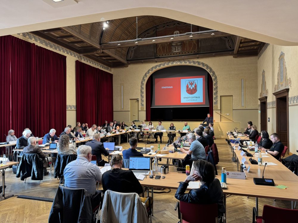 Im Saal des Stadthauses beraten viele Teilnehmende an Tischen; auf der Leinwand stehen Wappen und Stadt Cottbus/Chóśebuz.