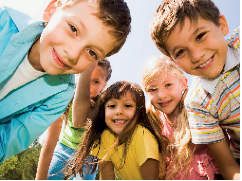 Mehrere Kinder lachen gemeinsam im Freien und schauen neugierig in die Kamera, während sie sich über die Wiese beugen und dicht zusammenstehen.
